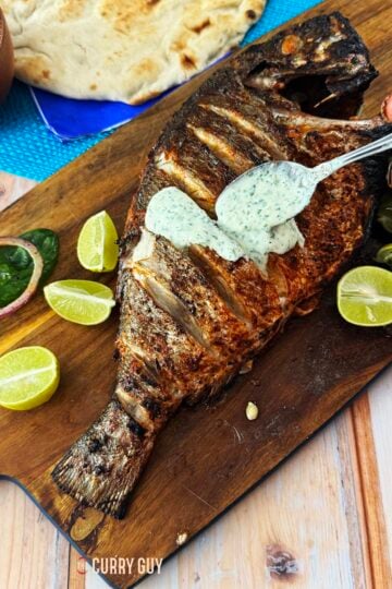 grilled whole fish with raita on a serving board with naans and salad.