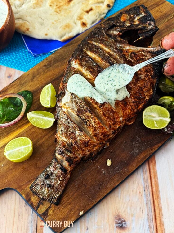 grilled whole fish with raita on a serving board with naans and salad.