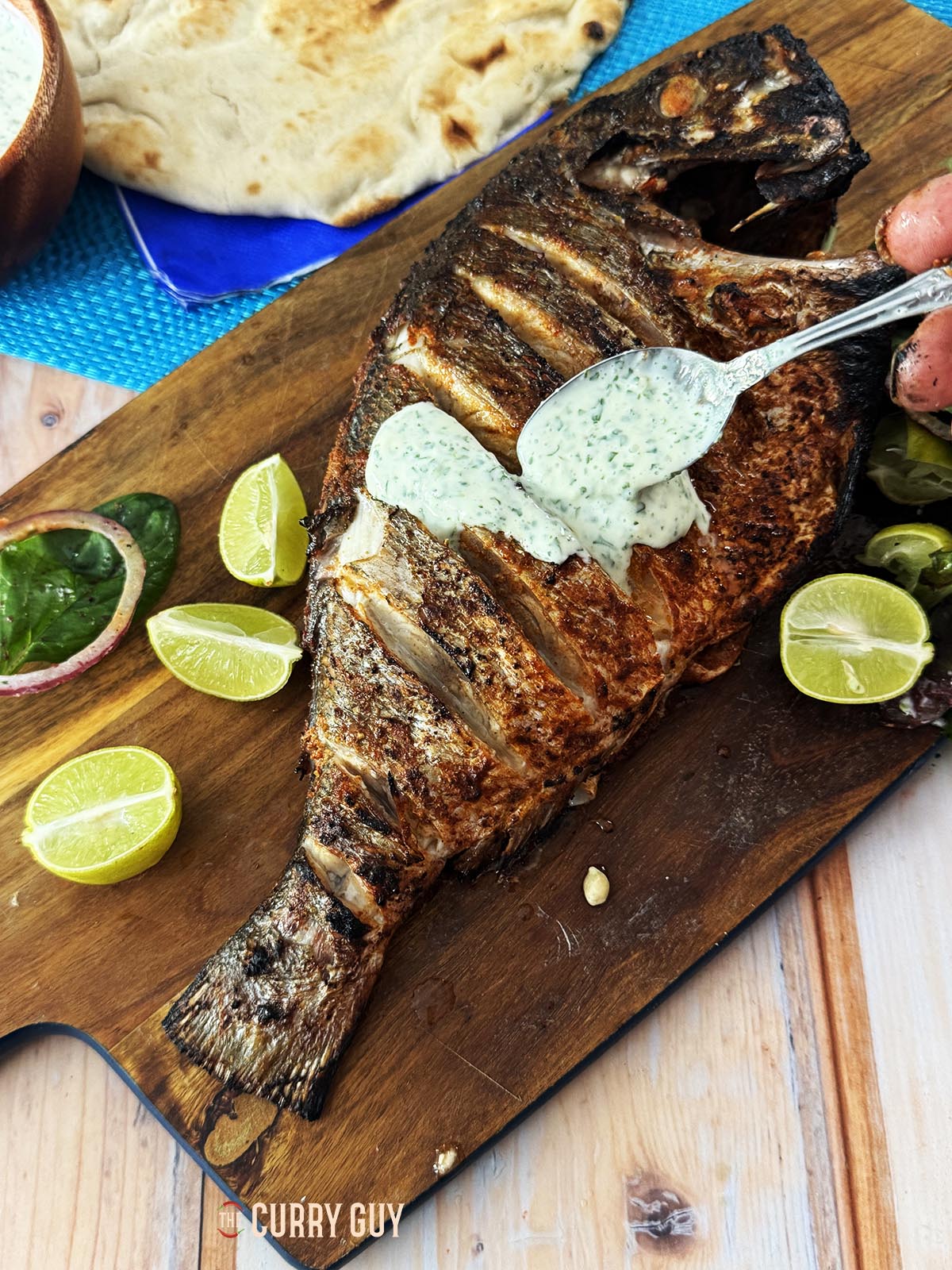 grilled whole fish with raita on a serving board with naans and salad.