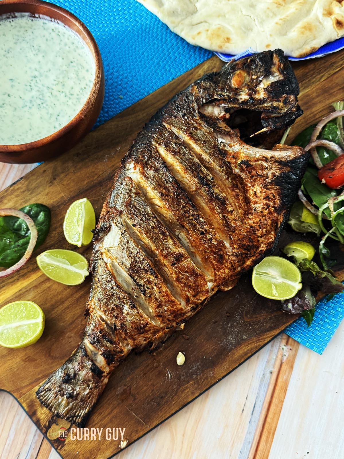 Grilled whole fish on a serving platter with raita, limes, salad and naans.