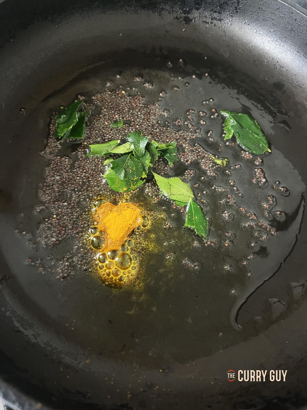 Infusing mustard seeds, curry leaves and ground turmeric into oil in a pan.