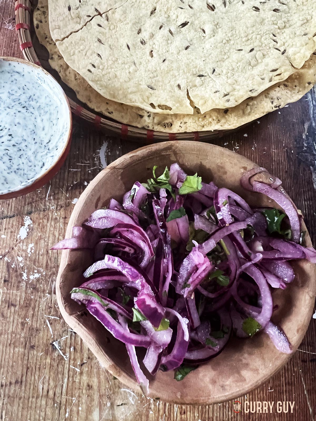 Spicy red onion pickle in a serving bowl next to a raita and poppadoms.