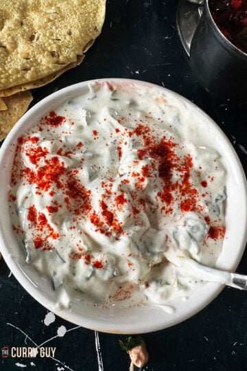 Cucumber raita in a serving bowl garnished with chilli powder.