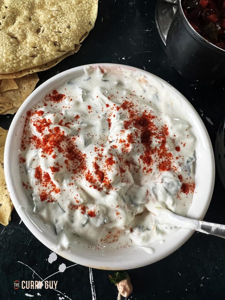 Cucumber raita in a serving bowl garnished with chilli powder.