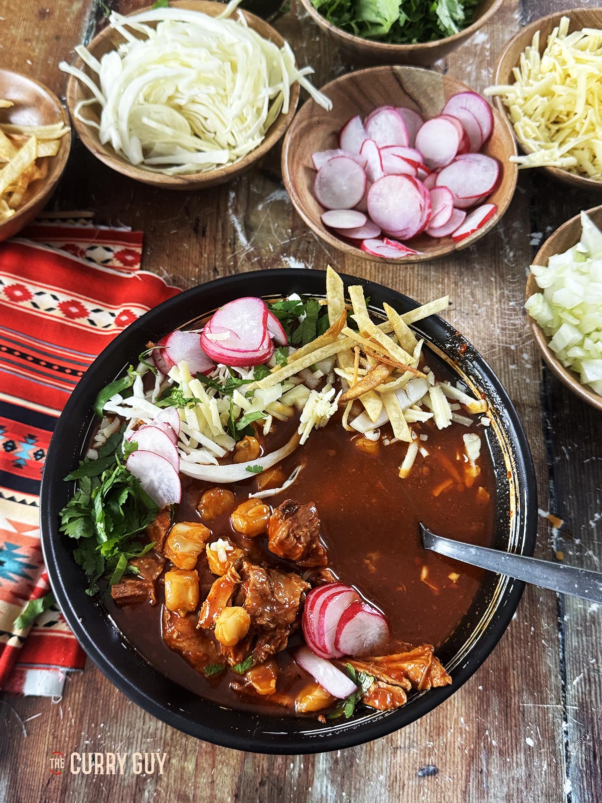 A bowl of pozole rojo served with garnishes such as cheese, shredded cabbage, sliced radishes, coriander (cilantro) and crispy corn chips.