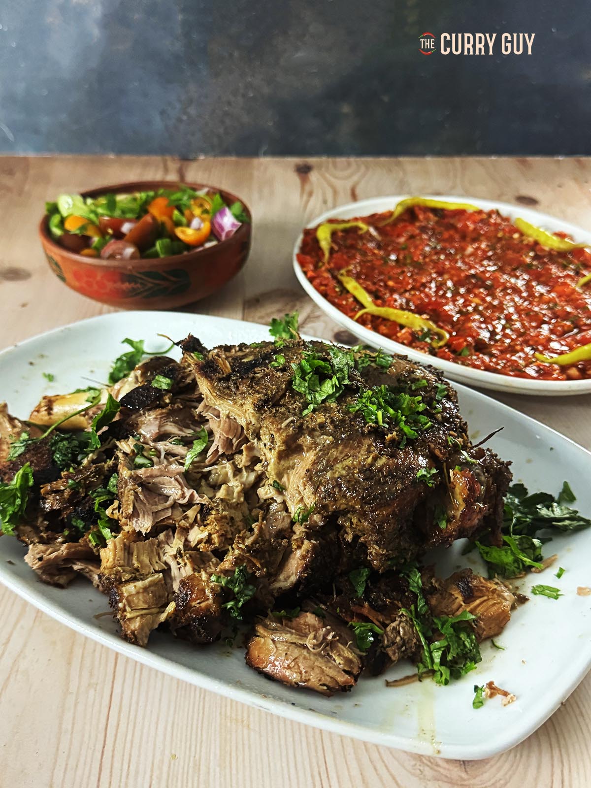 Slow cooked lamb leg on a serving platter with additional side salads in the background.