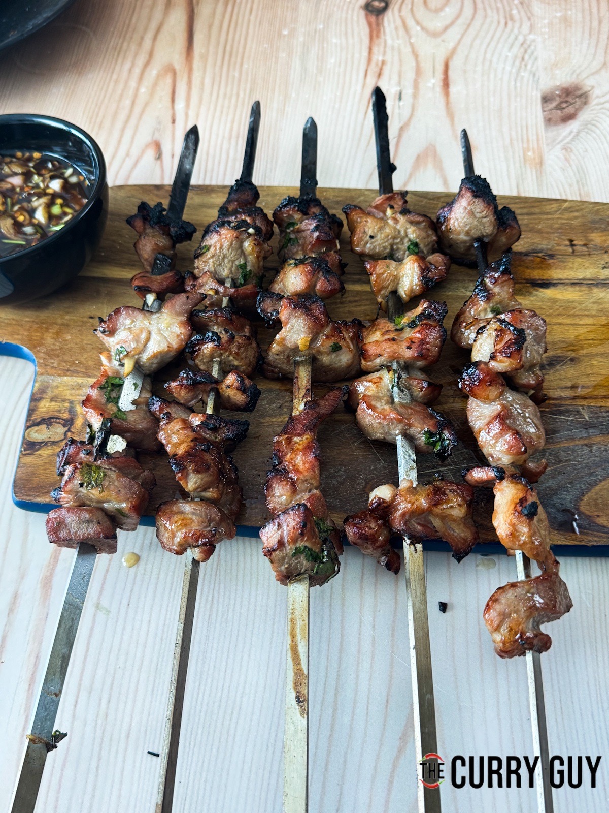 The pork skewers resting on a serving board next to nam Jim jaew sauce.